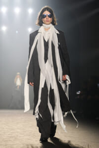 Model on a runway wearing a black pinstripe suit with a long white scarf and sunglasses, making a bold fashion statement.
