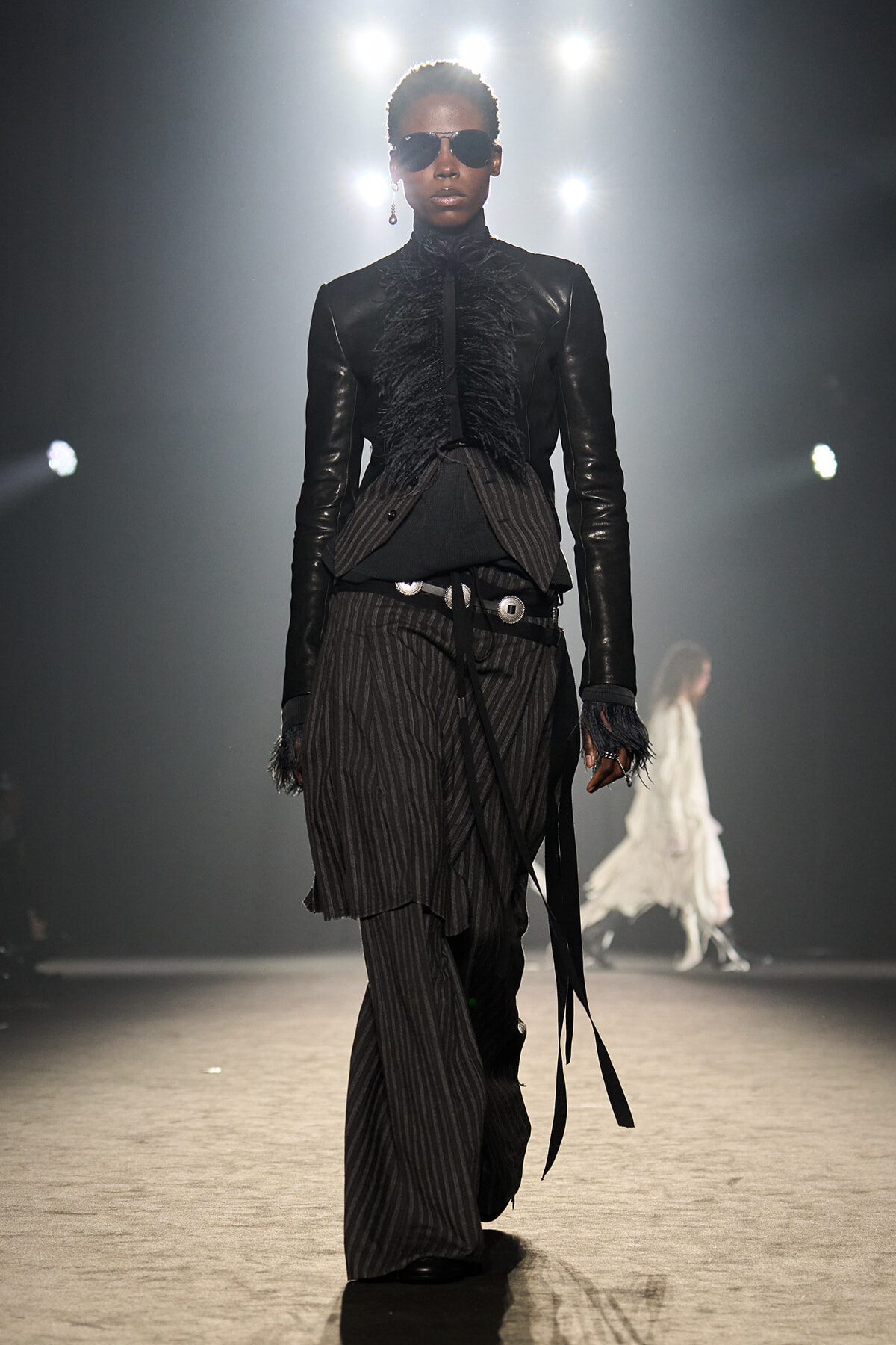 Model walking a runway in a black leather blazer with feathered front and pinstripe wide-leg pants, wearing sunglasses.