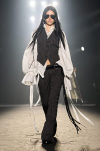 Model wearing a pinstripe vest and pants with an oversized white shirt on a runway, sunglasses on, walking forward confidently.