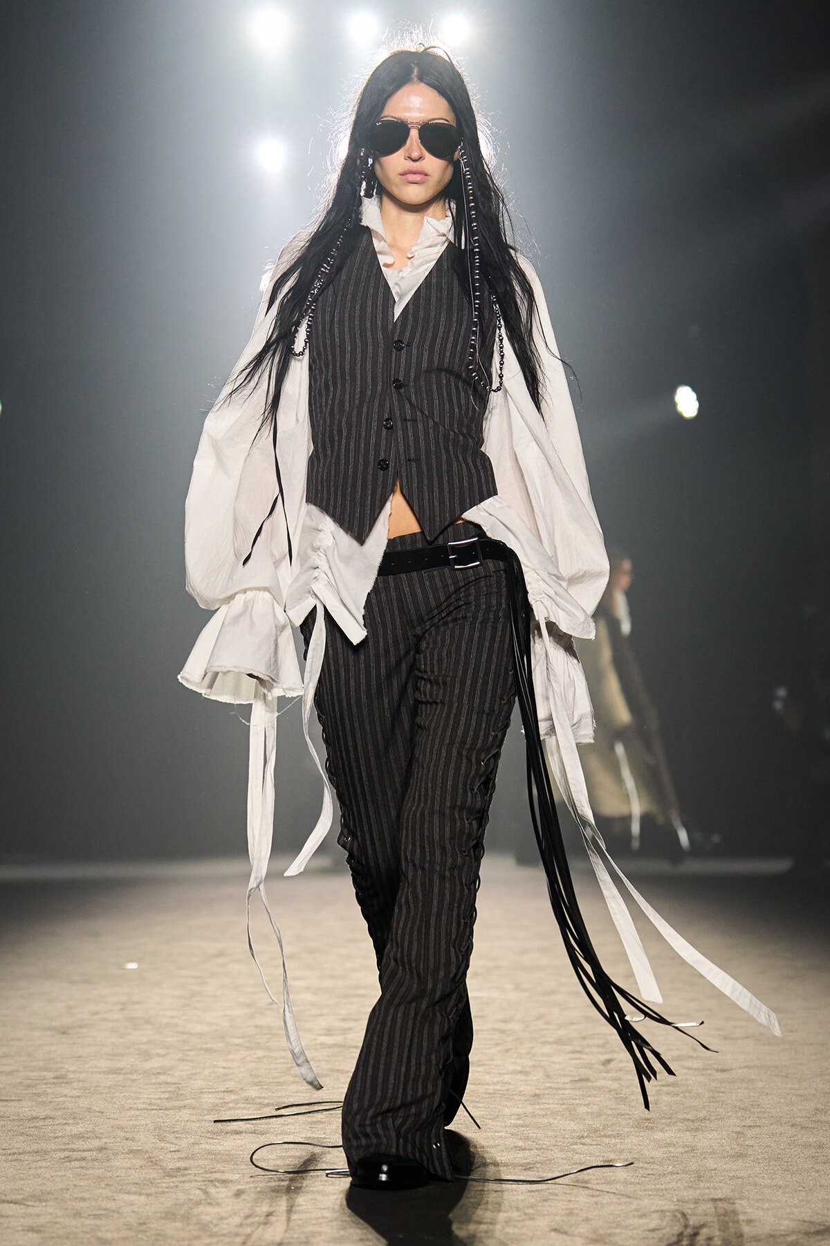 Model wearing a pinstripe vest and pants with an oversized white shirt on a runway, sunglasses on, walking forward confidently.