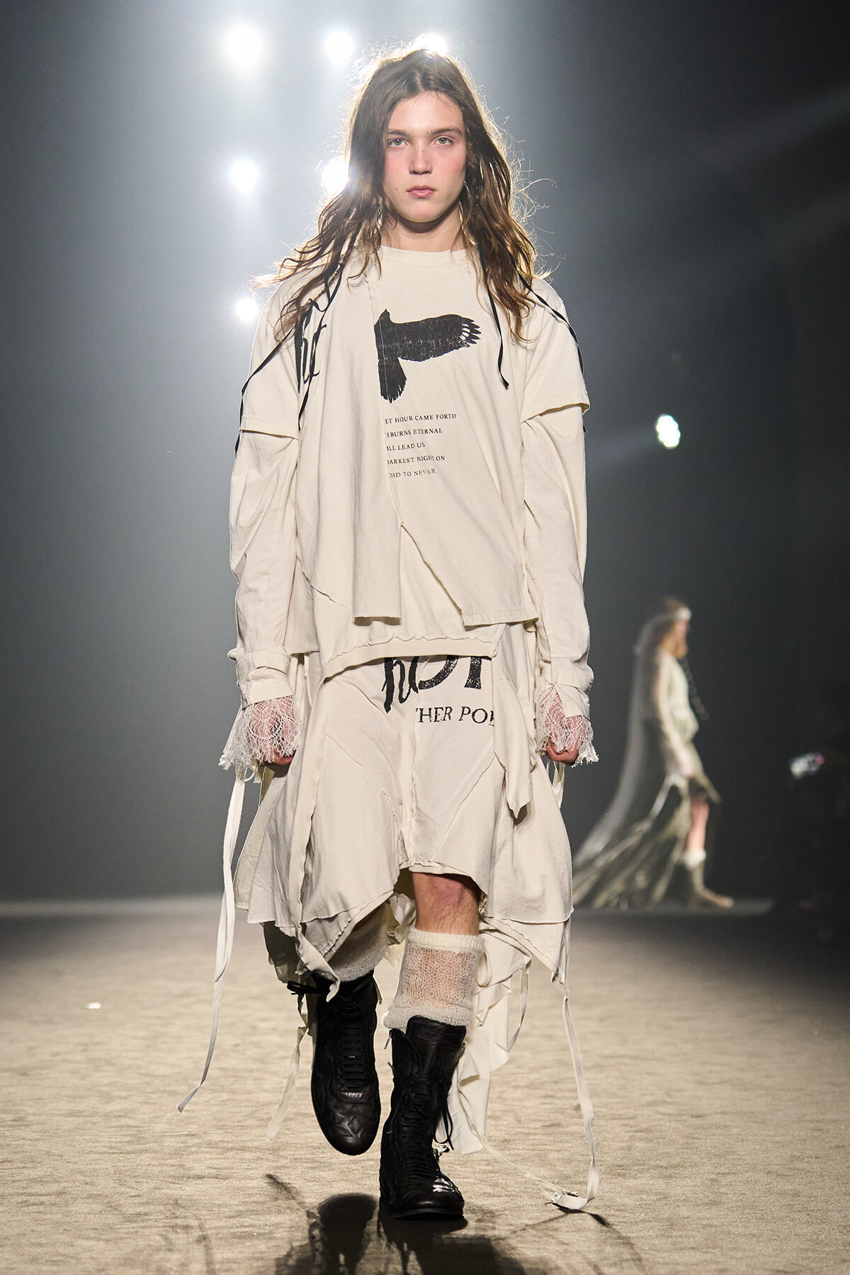 Model on a fashion runway wearing a loose, cream-toned layered outfit with long sleeves, lace cuffs, and black combat boots under bright stage lights.
