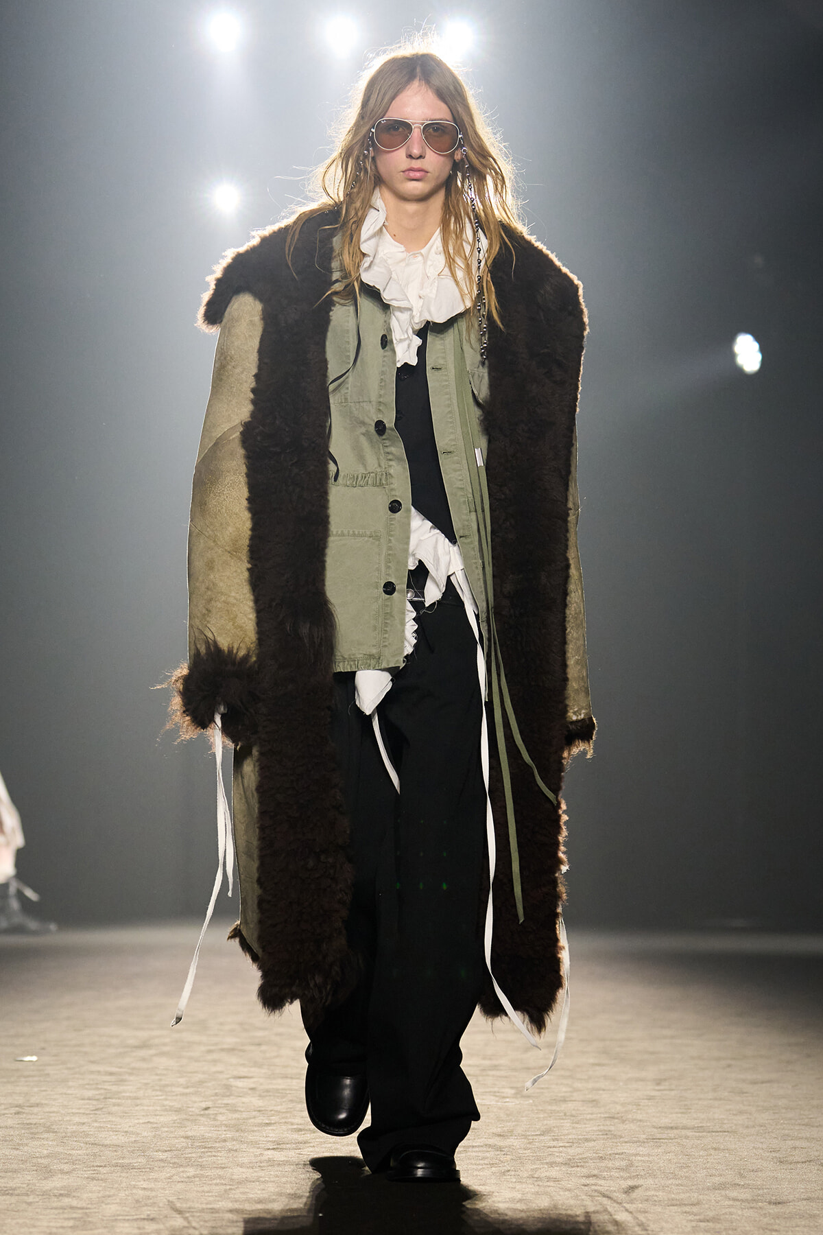 Model walking a runway in a long brown shearling coat over an olive-green jacket, white ruffled blouse, and black pants with sunglasses.
