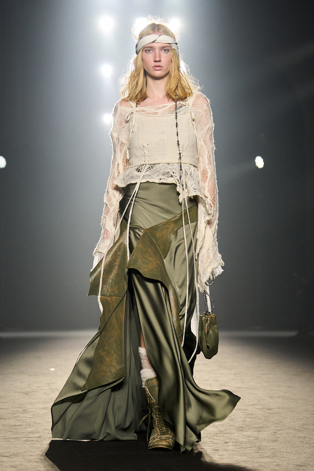 Model walks a runway in a boho-inspired cream lace top layered over a cropped cream corset, olive-green skirt, and tan lace-up boots, with a small olive bag and white headband under bright backstage lights.