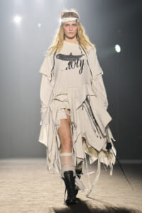 Runway model in oversized off-white layered outfit with a graphic tee, white headband, and black boots walking a fashion show runway.