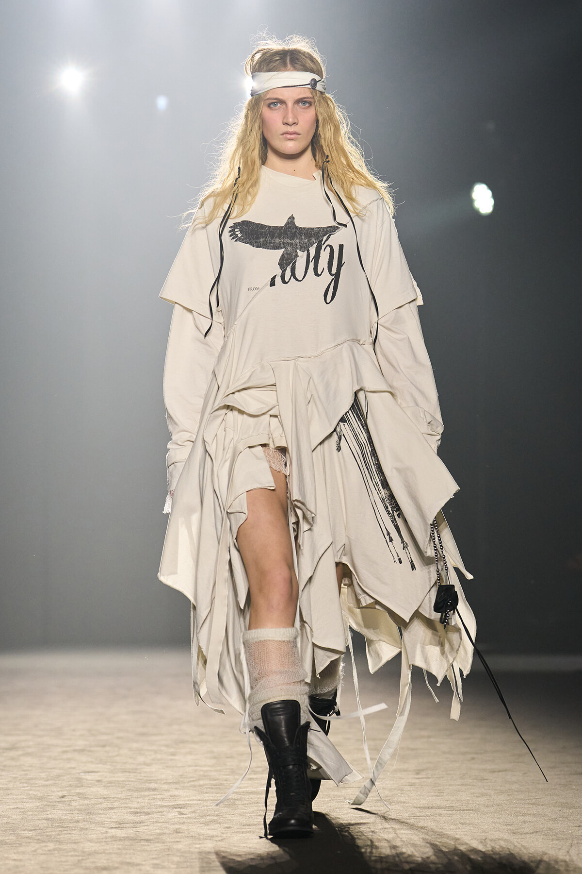 Runway model in oversized off-white layered outfit with a graphic tee, white headband, and black boots walking a fashion show runway.