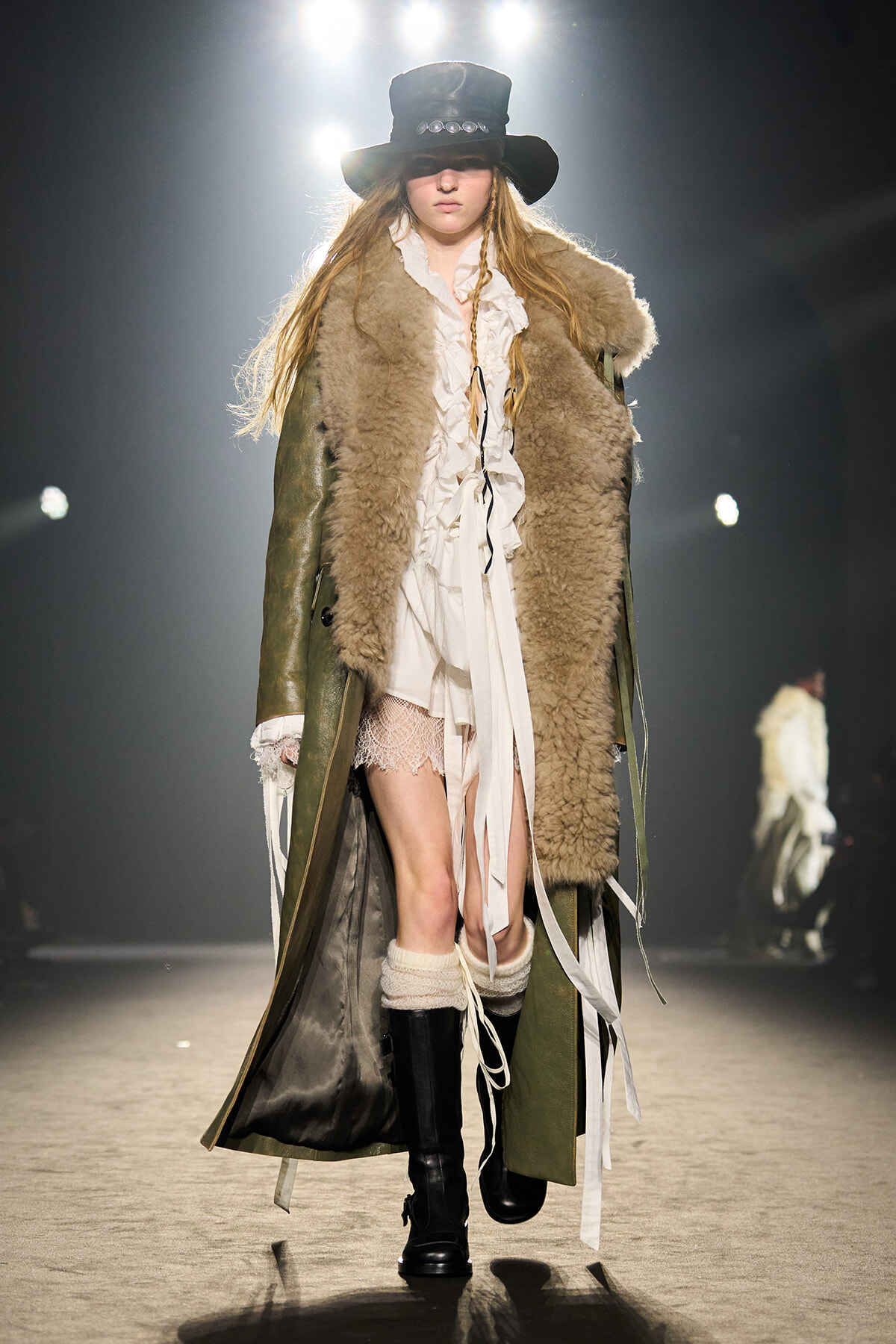 Model on a runway wearing an olive shearling coat over a white ruffled dress, with black boots and a wide-brim hat