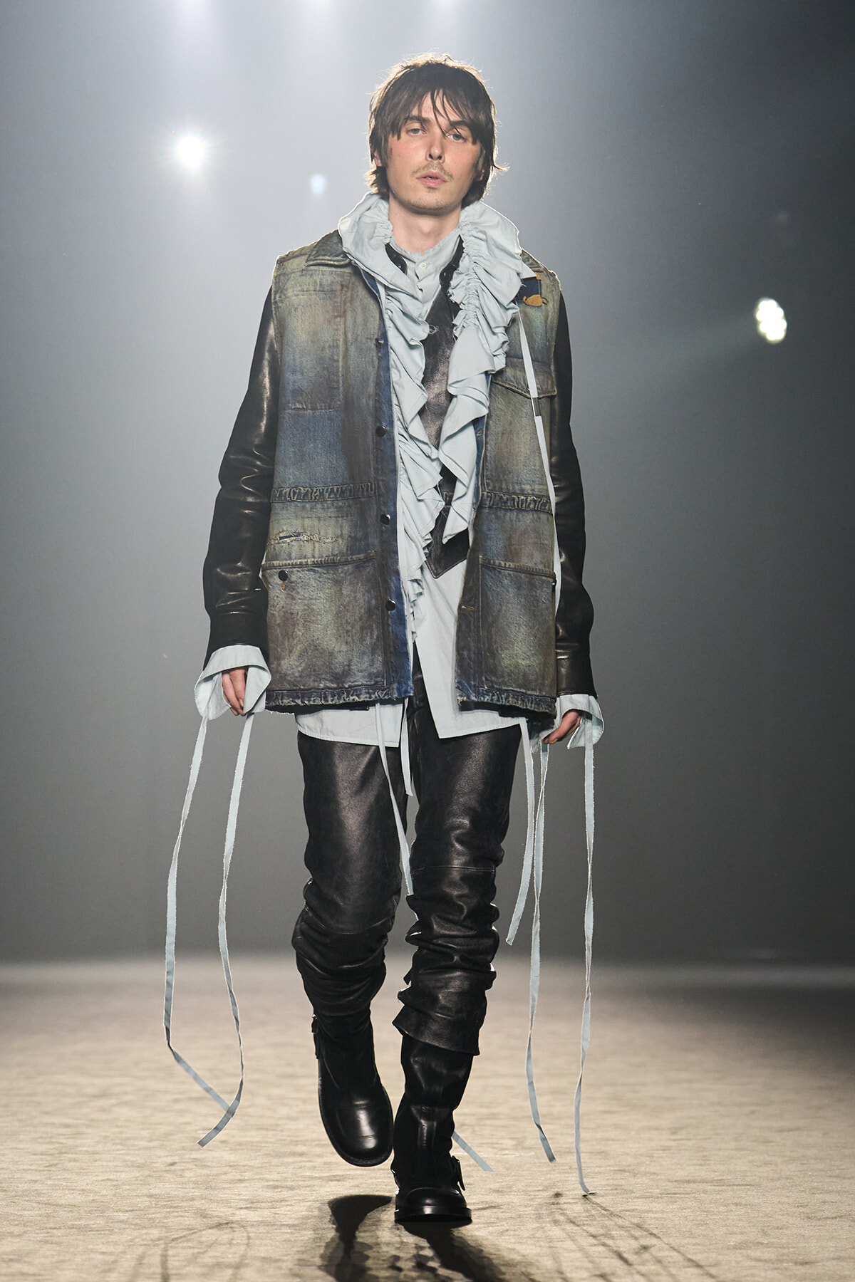 Male model on a runway wearing a distressed denim vest over a light-gray ruffled shirt, black leather pants, and tall black boots with long white ribbons at the sleeves.