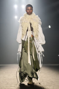 Model walks the runway in a voluminous white shirt with a cream fur scarf and a layered olive-green skirt under bright spotlights.