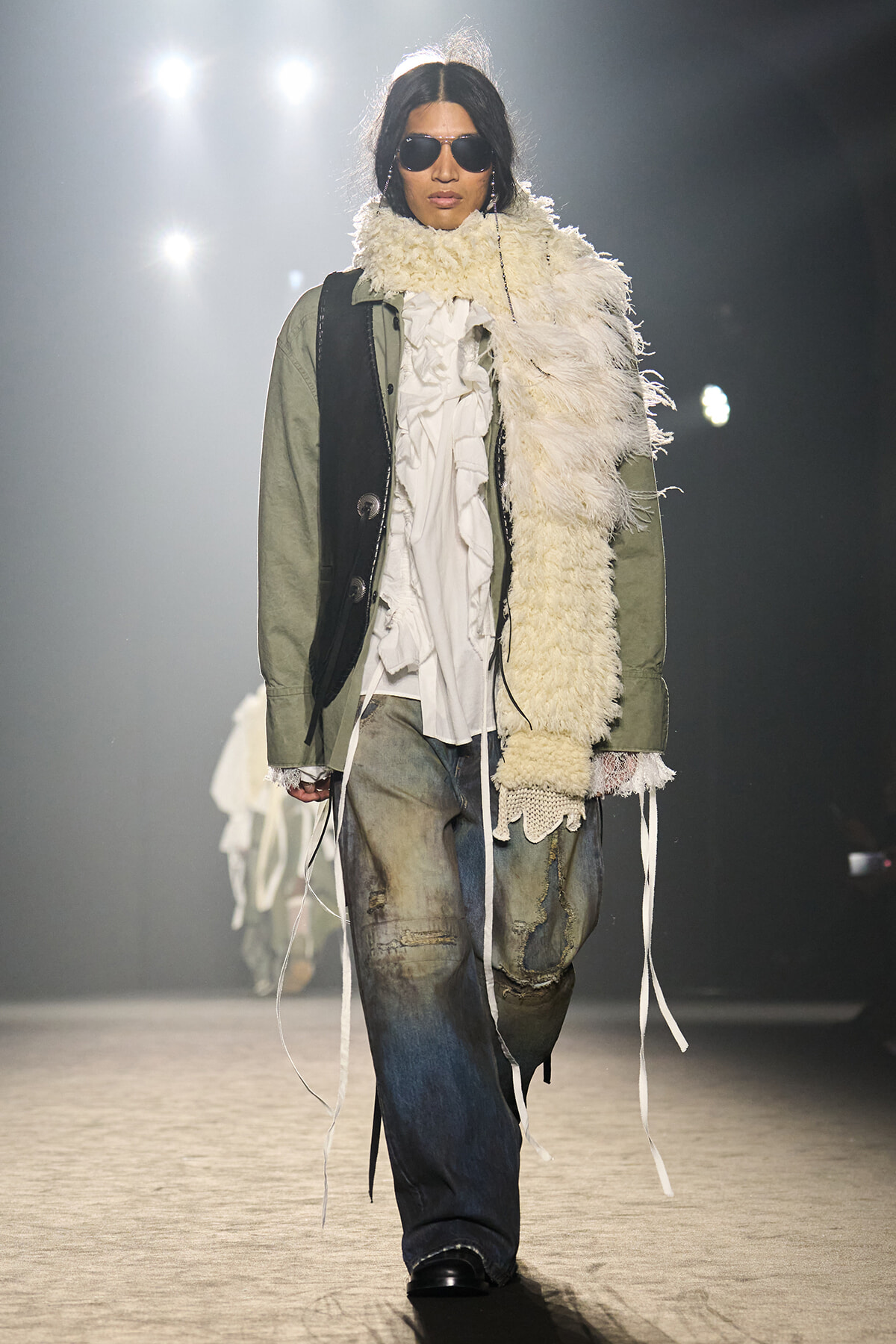 Runway model wearing an olive-green jacket, white ruffled shirt, cream shearling scarf, distressed jeans, and sunglasses.