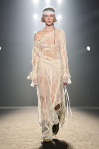 Model walks a fashion show in a sheer cream lace dress with an off-shoulder neckline and white headband under bright runway lights.