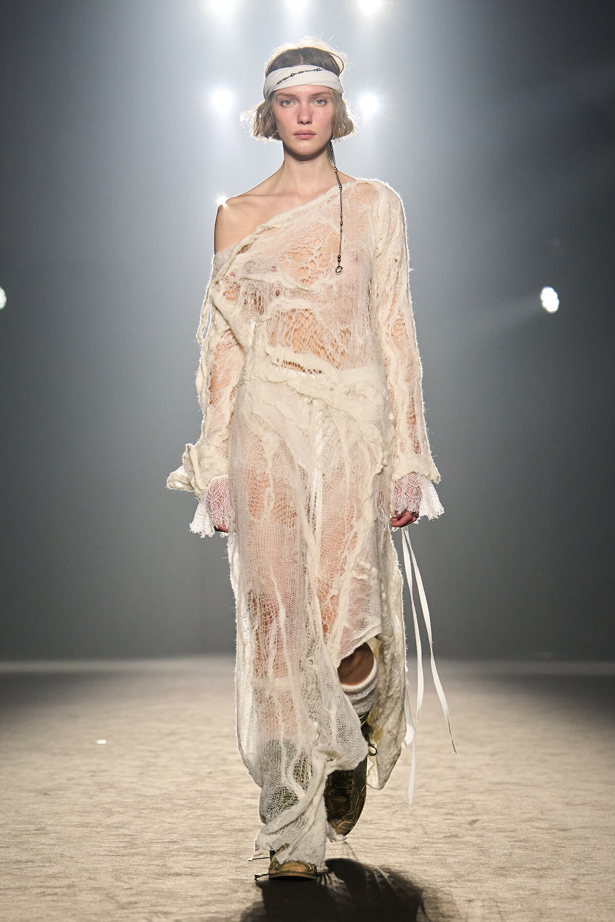 Model walks a fashion show in a sheer cream lace dress with an off-shoulder neckline and white headband under bright runway lights.