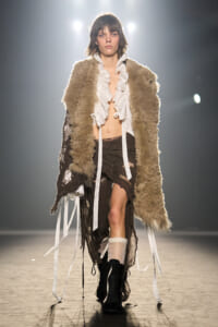 Model walking the runway in a shaggy beige fur coat over a white ruffled blouse and distressed brown skirt, with black boots in view.