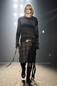 Model walking a runway in a black and gray striped knit sweater, layered plaid skirt with black fabric, and tall black lace-up boots, wearing dark sunglasses under bright backstage lights.