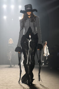 Model walks the runway in a Gothic-inspired look: wide-brimmed black hat, gray tailored jacket, and black leather pants with hanging ribbons. The ensemble is monochrome with dramatic, elongated sleeves and fringe details on the sides.