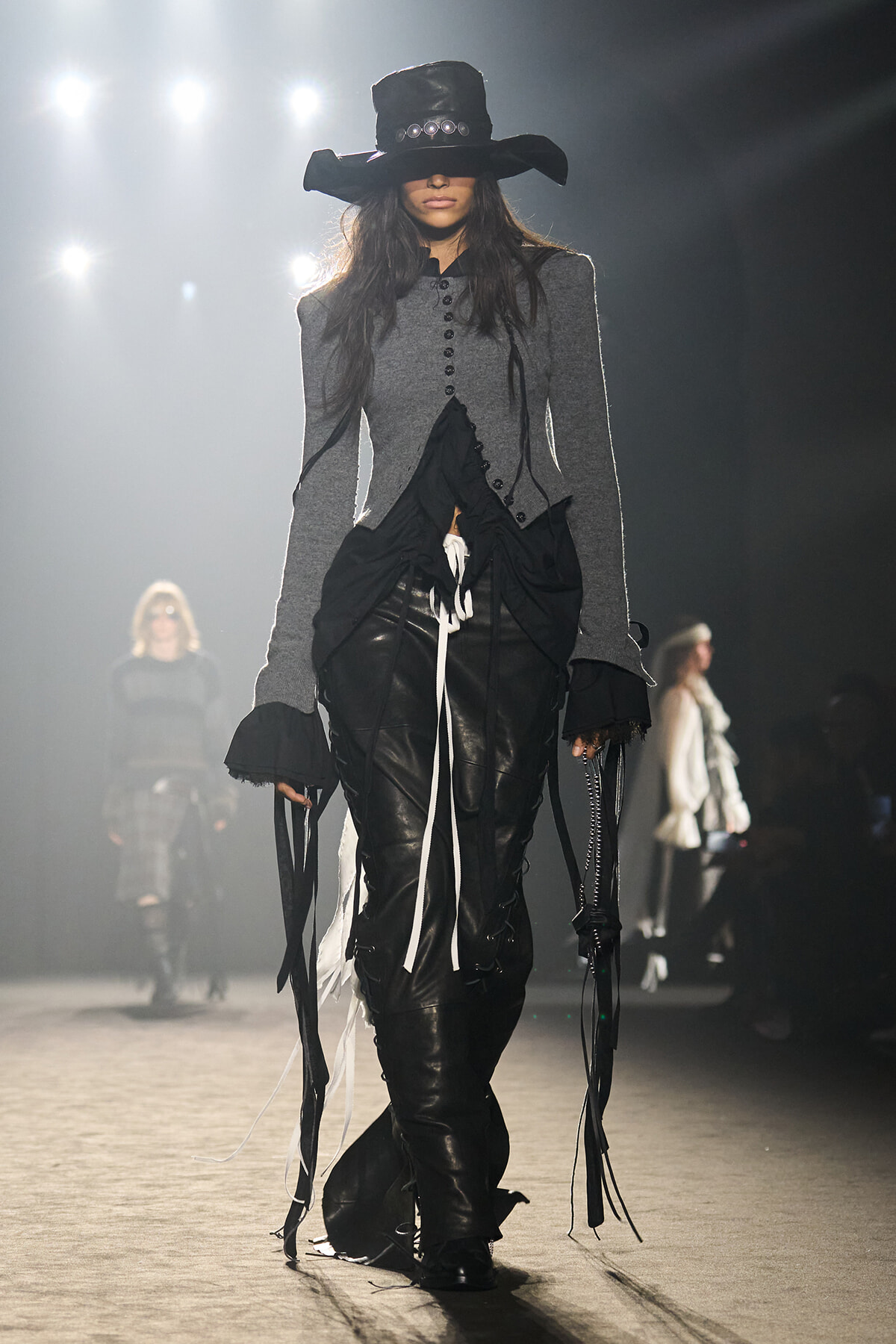 Model walks the runway in a Gothic-inspired look: wide-brimmed black hat, gray tailored jacket, and black leather pants with hanging ribbons. The ensemble is monochrome with dramatic, elongated sleeves and fringe details on the sides.