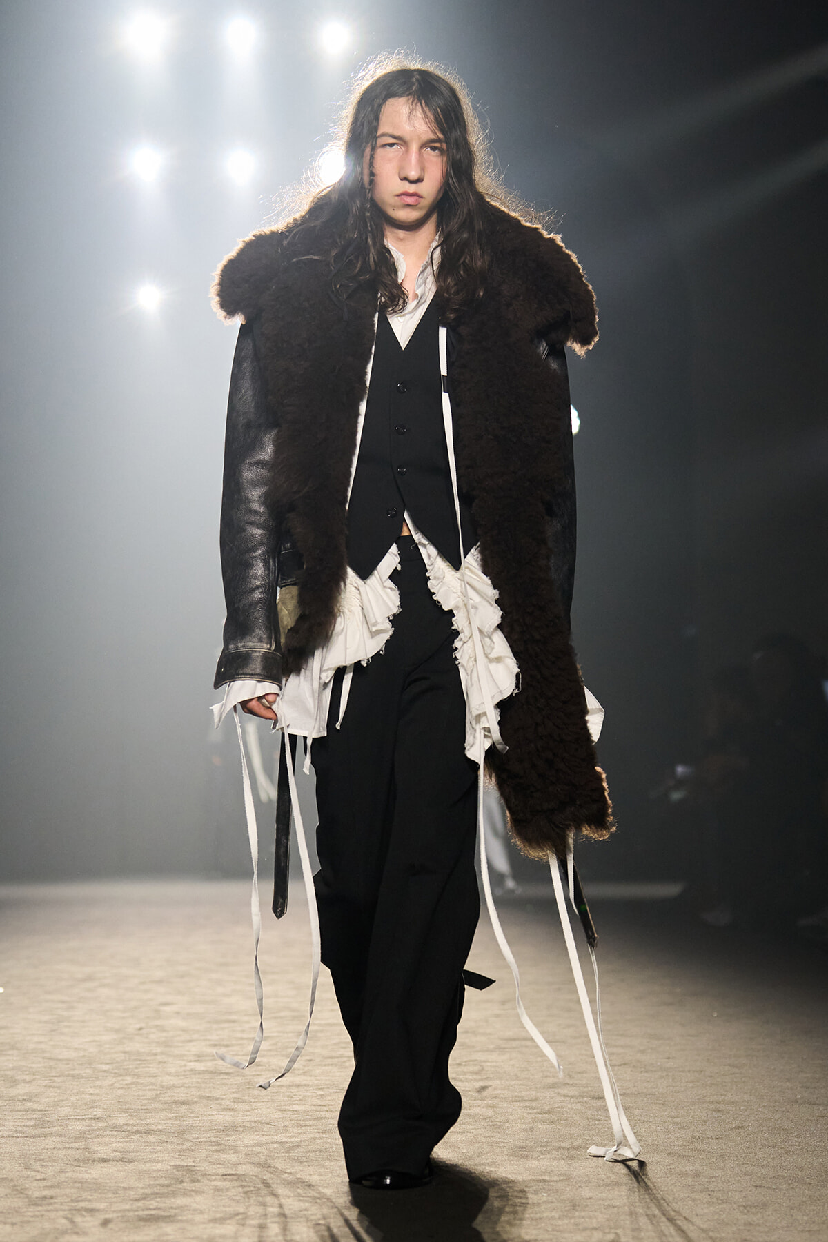 Model walking the runway in a dark fur coat over a black vest and white ruffled shirt.