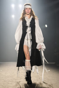 Model walking a runway in a white billowy blouse with lace cuffs, black vest overcoat, white headband, and chunky boots with leg warmerssueded for a bohemian look.