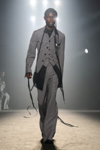 Male model walks runway in a grey pinstriped three-piece suit with black ribbons draped from shoulders and waistcoat.