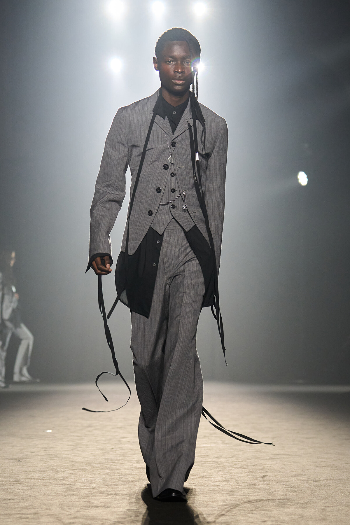 Male model walks runway in a grey pinstriped three-piece suit with black ribbons draped from shoulders and waistcoat.