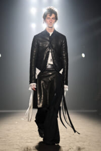 Male model walks the runway in a black leather jacket with an asymmetric hem and long fabric fringe over wide black pants with white underlayers.