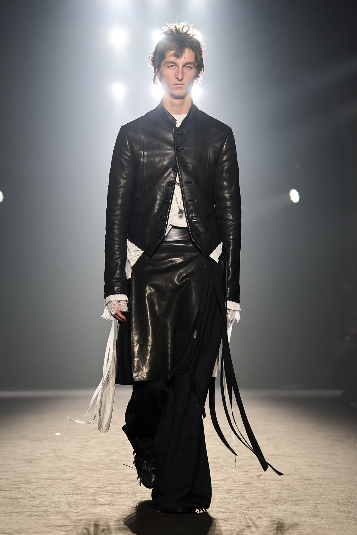 Male model walks the runway in a black leather jacket with an asymmetric hem and long fabric fringe over wide black pants with white underlayers.