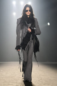 Model walking a fashion runway in a gray pinstripe suit with a black fringe scarf and sunglasses on.
