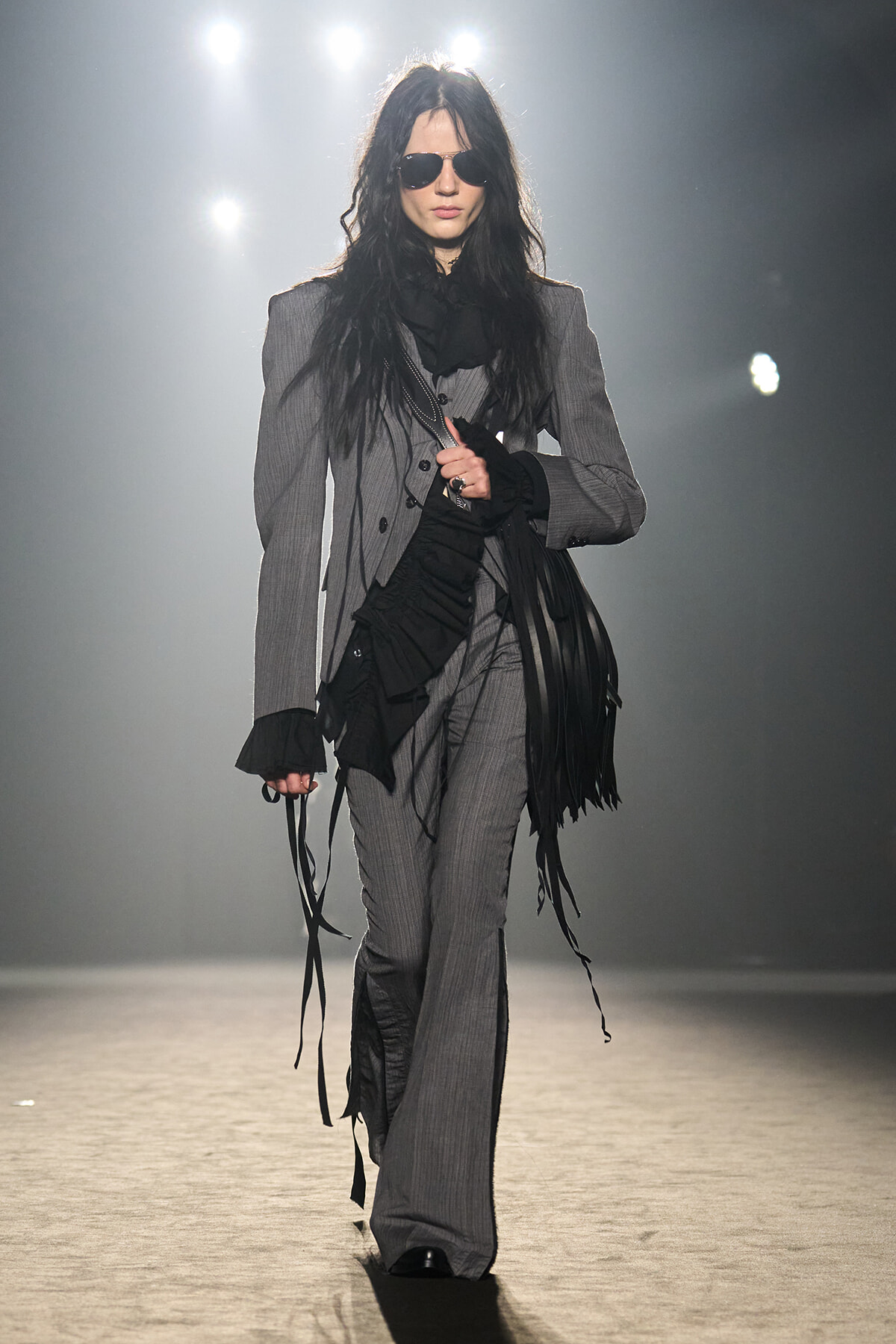 Model walking a fashion runway in a gray pinstripe suit with a black fringe scarf and sunglasses on.