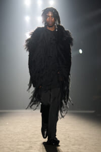 Female model on a fashion runway wearing a voluminous black feathered coat over a black blouse and pants, under bright stage lights.