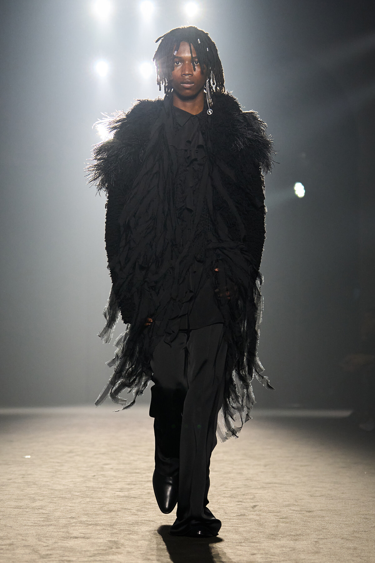 Female model on a fashion runway wearing a voluminous black feathered coat over a black blouse and pants, under bright stage lights.