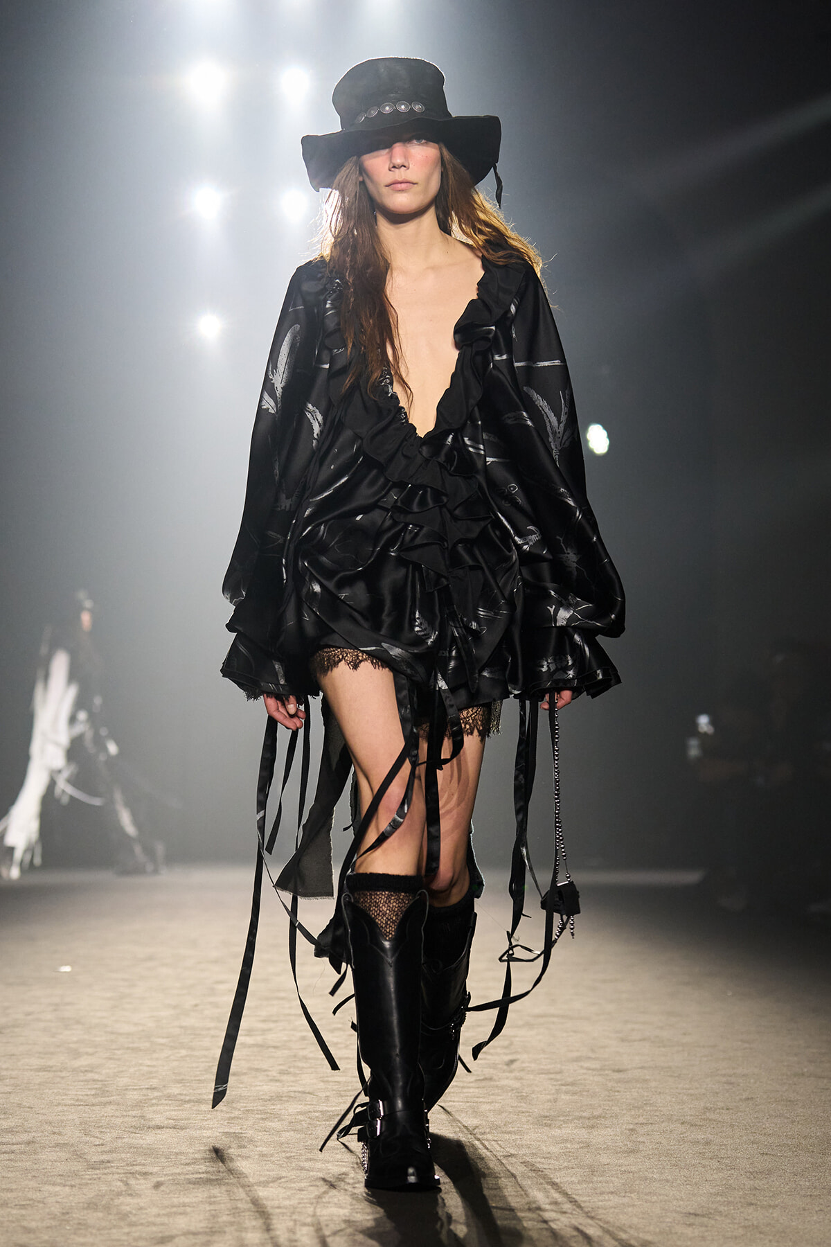 Model walks a runway in an all-black, ruffled satin dress with fringe ribbons, paired with a wide-brimmed black hat and knee-high boots.