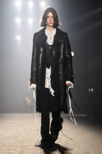 Model on runway wearing a long black leather coat over a white ruffled shirt, with black pants and lace-up details under bright backstage lights.