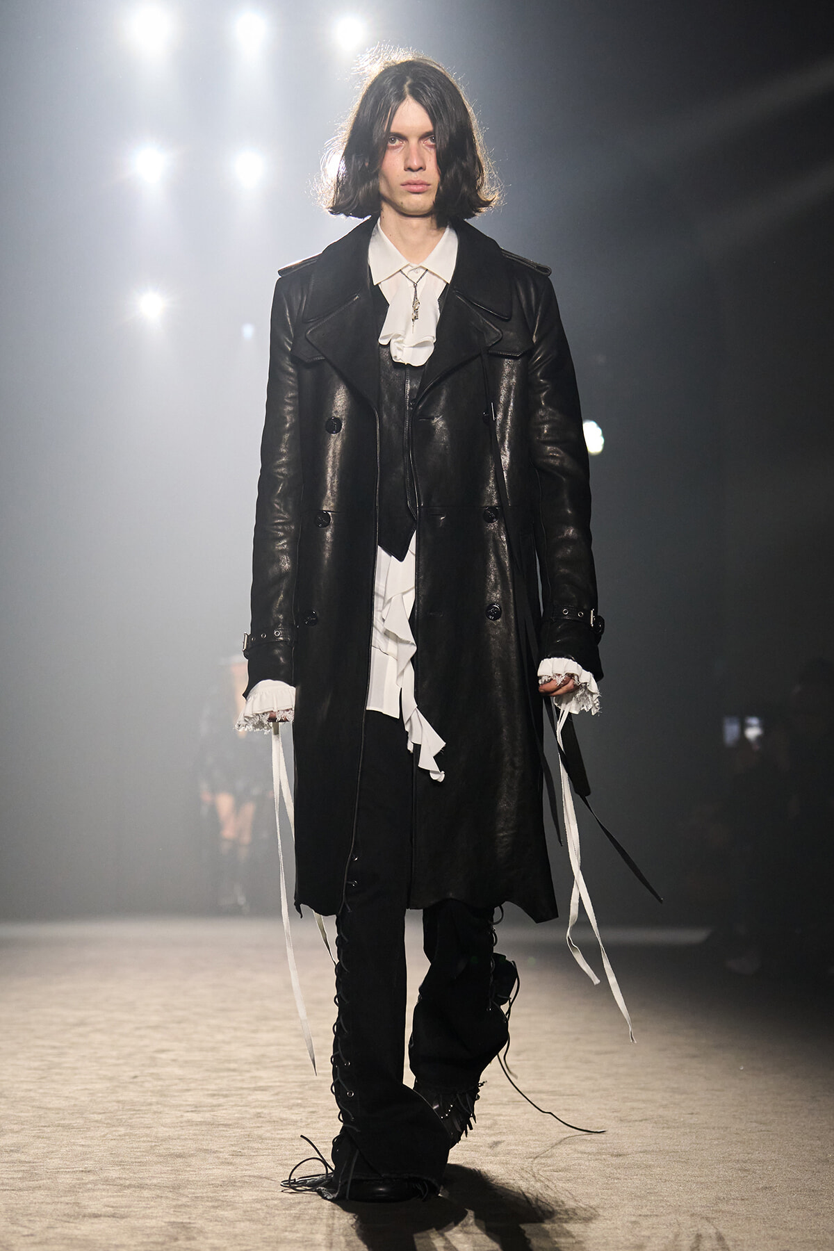 Model on runway wearing a long black leather coat over a white ruffled shirt, with black pants and lace-up details under bright backstage lights.