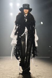 Model on a runway wearing a black wide-brim hat, long black fringe vest, white voluminous sleeves, and black leather pants.