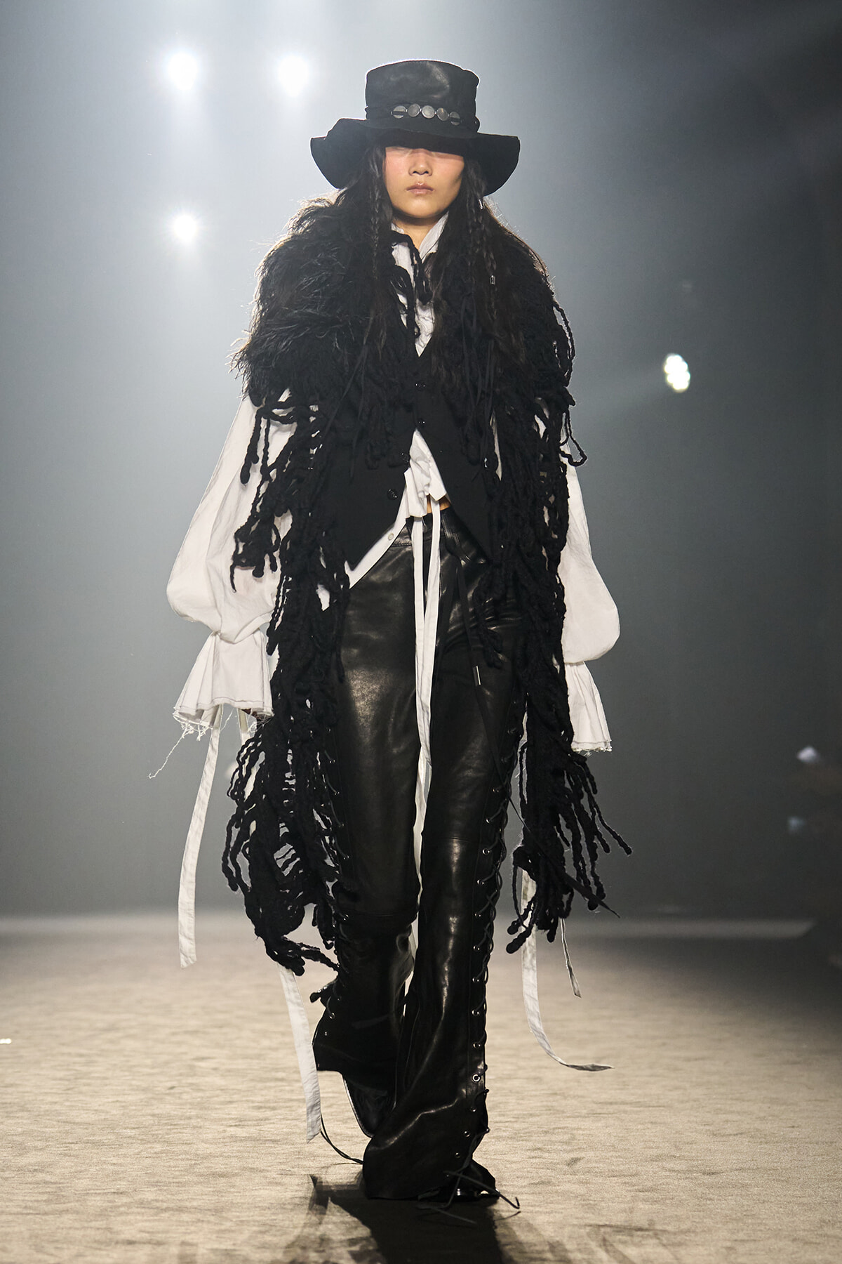 Model on a runway wearing a black wide-brim hat, long black fringe vest, white voluminous sleeves, and black leather pants.