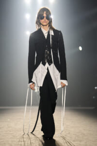 Model on runway wearing a black blazer, white shirt, black trousers, and long white ribbons hanging from sleeves.