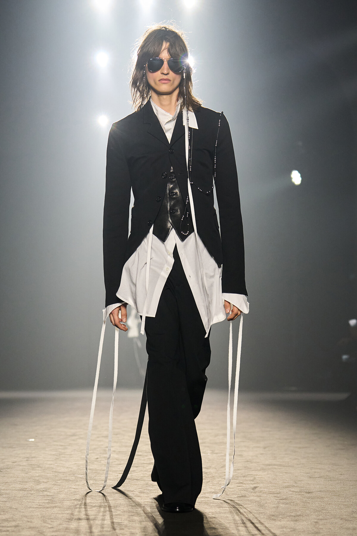 Model on runway wearing a black blazer, white shirt, black trousers, and long white ribbons hanging from sleeves.