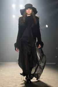 Model walks the runway in a textured black fur-like jacket, wide-brim hat, and flowing sheer black skirt with ankle boots.