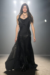 Female model walking a dim runway in a long black layered gown with a draped neckline and metallic accessories, spotlight behind her.