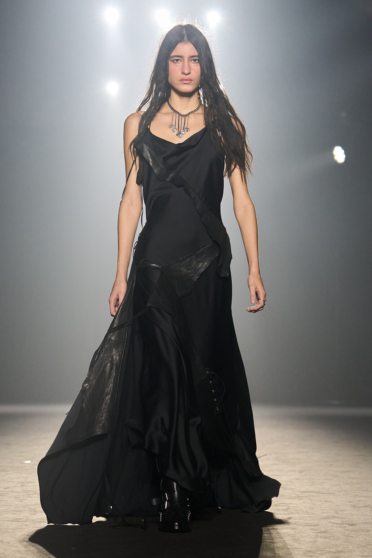 Female model walking a dim runway in a long black layered gown with a draped neckline and metallic accessories, spotlight behind her.