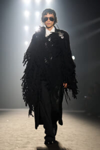 Model on a runway wearing a large black fringed cape, white shirt, black pants, and sunglasses, illuminated by bright stage lights behind.