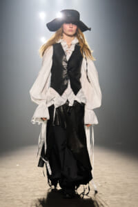 Model wearing a wide-brimmed black hat, white ruffled blouse, black vest, and tiered black skirt on a runway.