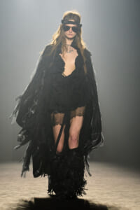 Model walks a runway in a dramatic black, ruffled cape with a plunging neckline and lace hem, paired with fringed boots and sheer stockings.
