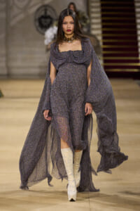 Model on a runway wearing a sheer, dark purple floral gown with cape sleeves and white patterned knee-high boots.