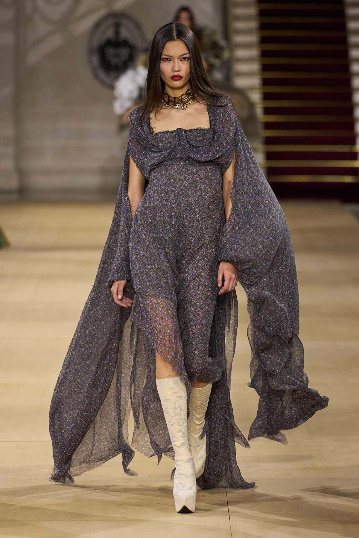 Model on a runway wearing a sheer, dark purple floral gown with cape sleeves and white patterned knee-high boots.