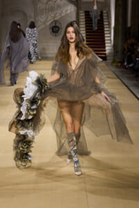 Model walking the runway in a sheer brown gown, holding a large flower bouquet, with patterned knee-high boots.