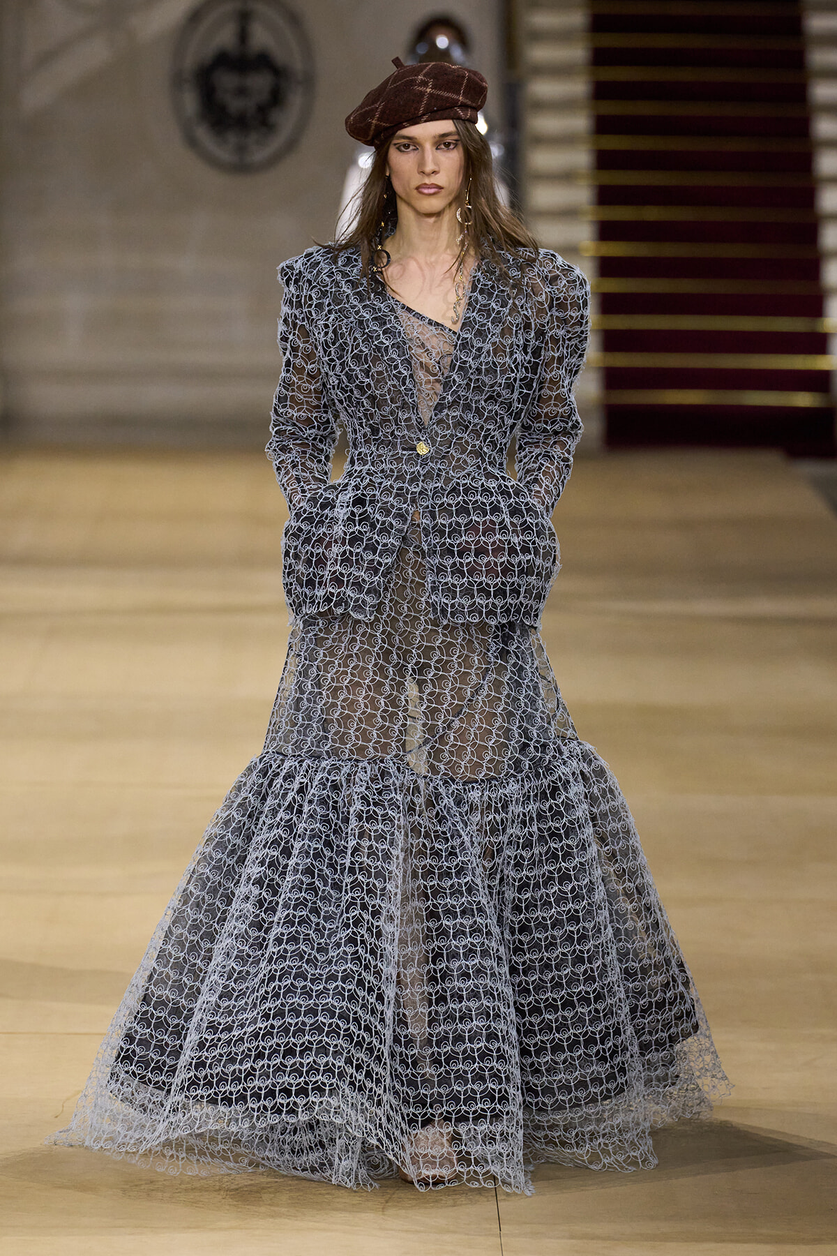 Model walks a fashion runway wearing a sheer gray lace suit with a matching tiered skirt and a brown beret, neutral makeup, minimal jewelry.