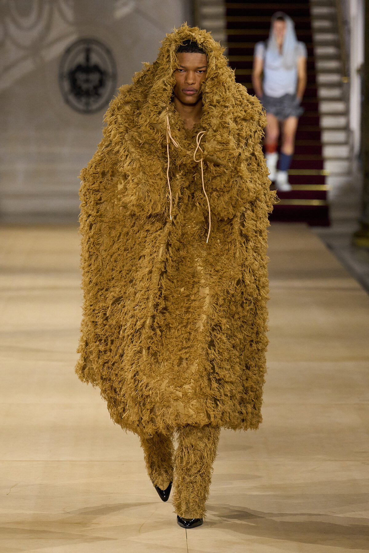 Model walking the runway in a full-length brown shaggy fur outfit with a hood and matching pants, filling the look from head to toe.