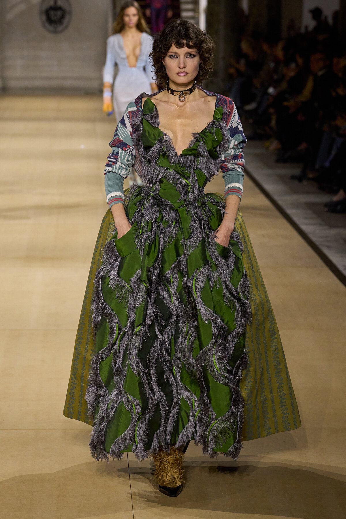 Model walks the runway in a green gown with dark gray feather-like fringe, layered over a patterned cardigan-sleeve top; choker necklace and audience in background.