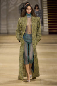 Model walking runway in a long, shimmering olive-green coat over a sheer teal-green bodysuit and pants ensemble, hands in pockets.