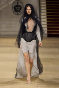 Runway model wearing a black veil over a sheer, lace-edged bodice with a beige, grid-pattern skirt and fishnet sleeves; long black cape-like overlay flows as she walks.